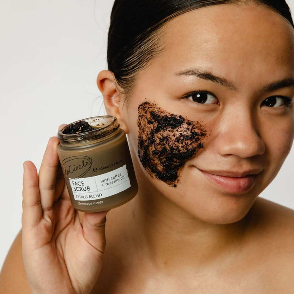 Woman applying coffee face scrub to her face with a jar of the product.