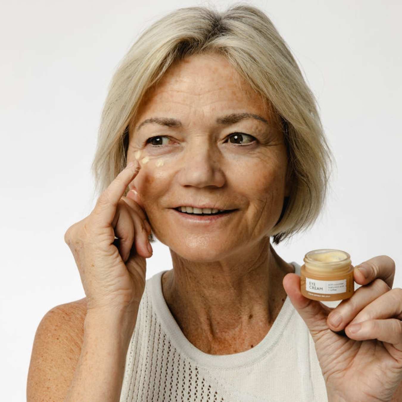 Woman applying cream to her face with a jar of cream in her hand on a white background