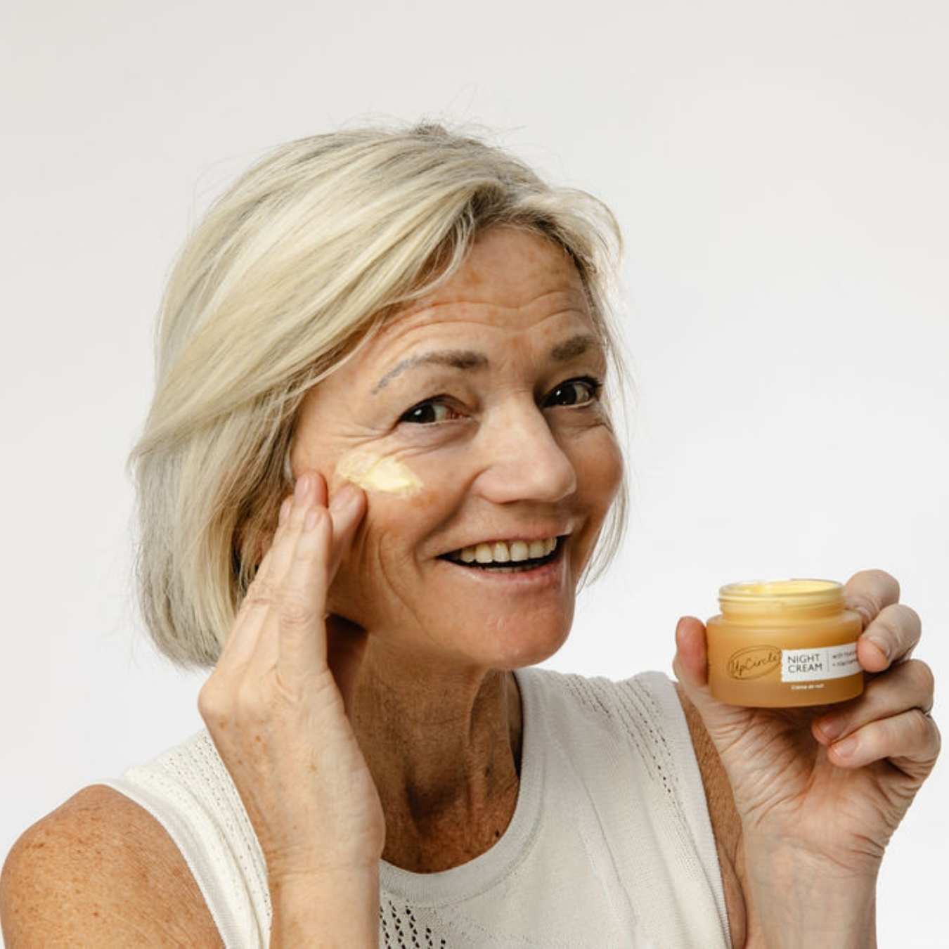 Woman applying cream to her face with a jar of cream in hand on a white background