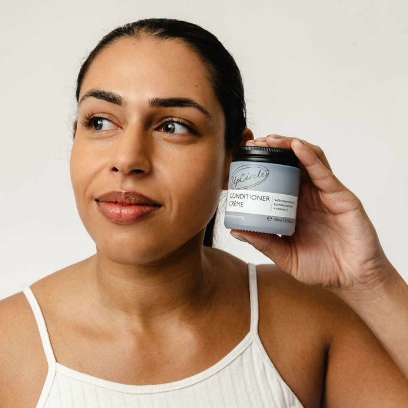 Woman holding a jar of UpCircle conditioner creme close to her ear against a plain background