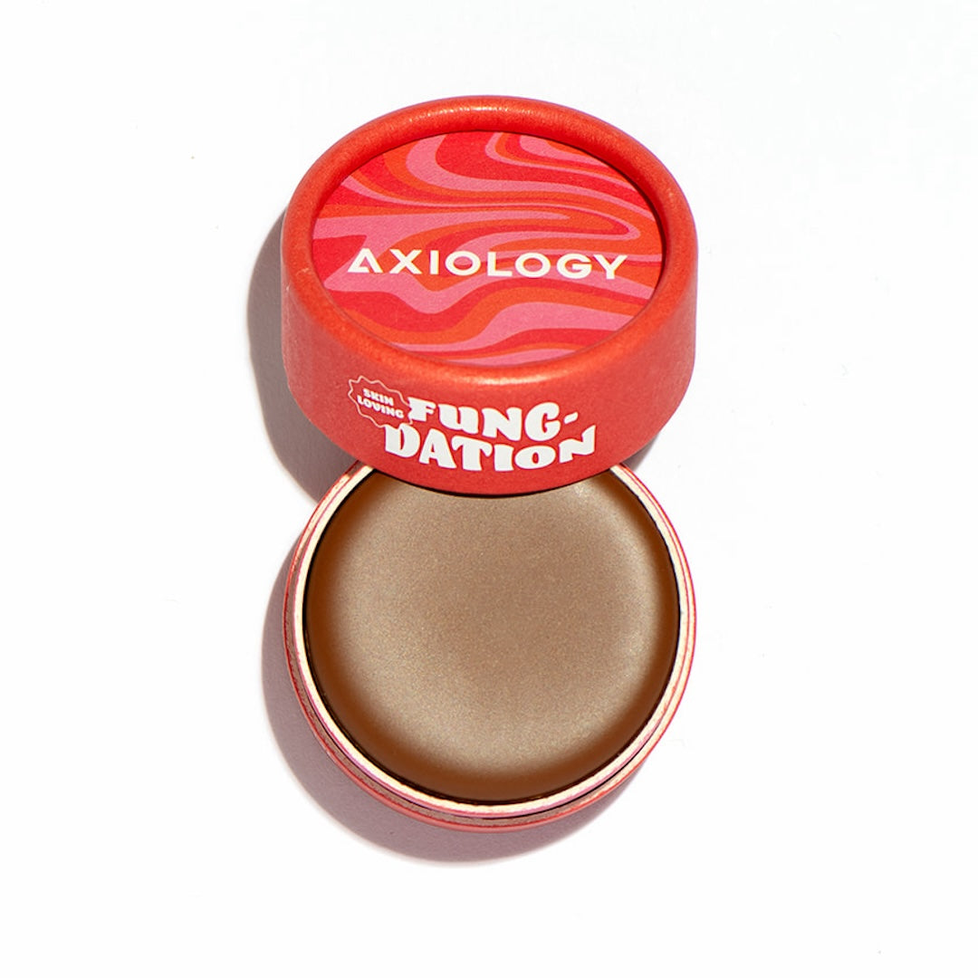 Open cosmetic pot with brown foundation and red lid featuring 'Axiology' branding on a white background.