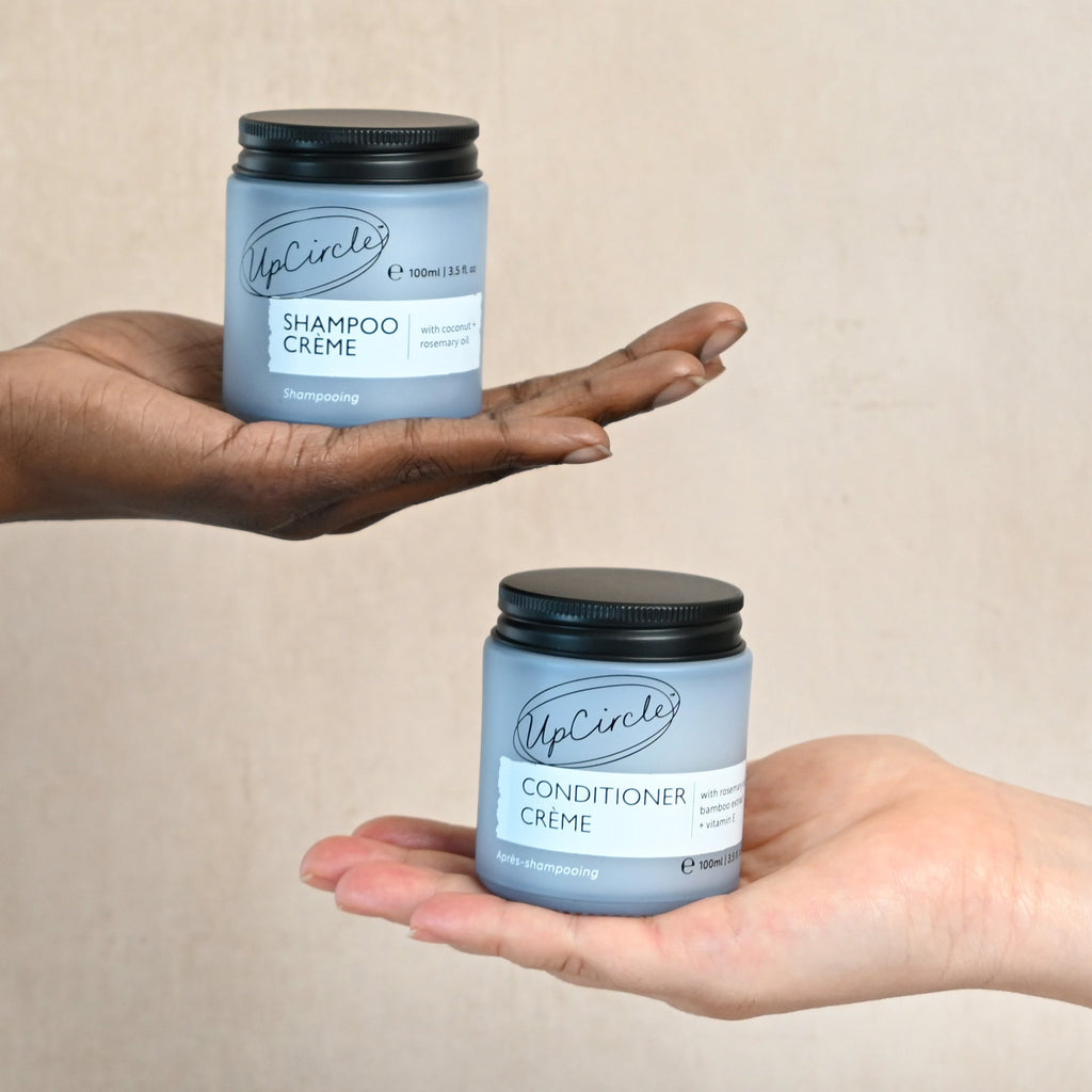 Two hands holding UpCircle's Shampoo Crème and Conditioner Crème jars against a beige background