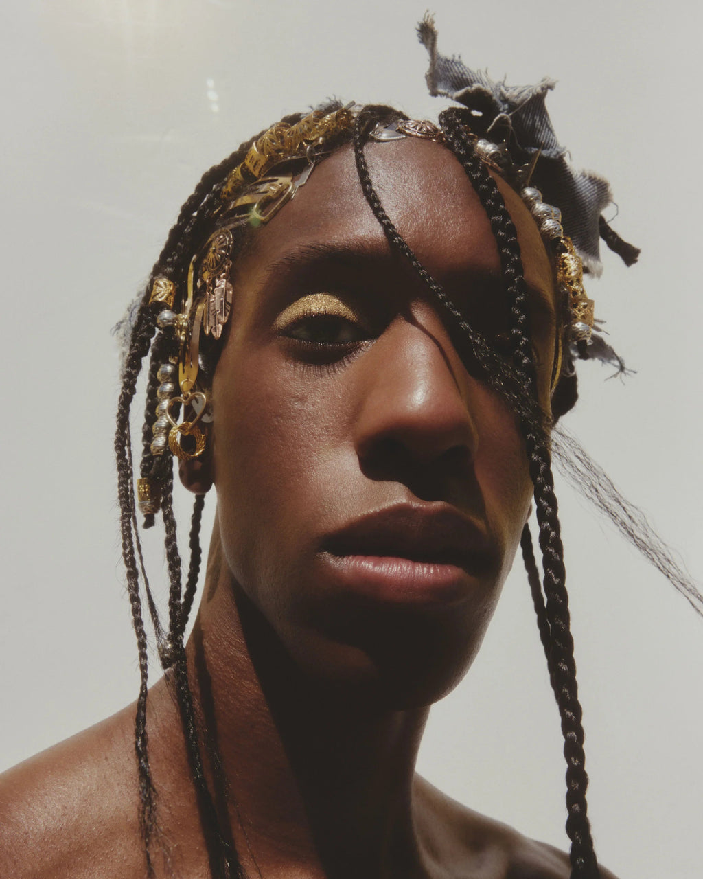 Person with braided hair wearing gold glitter eye makeup against a neutral background