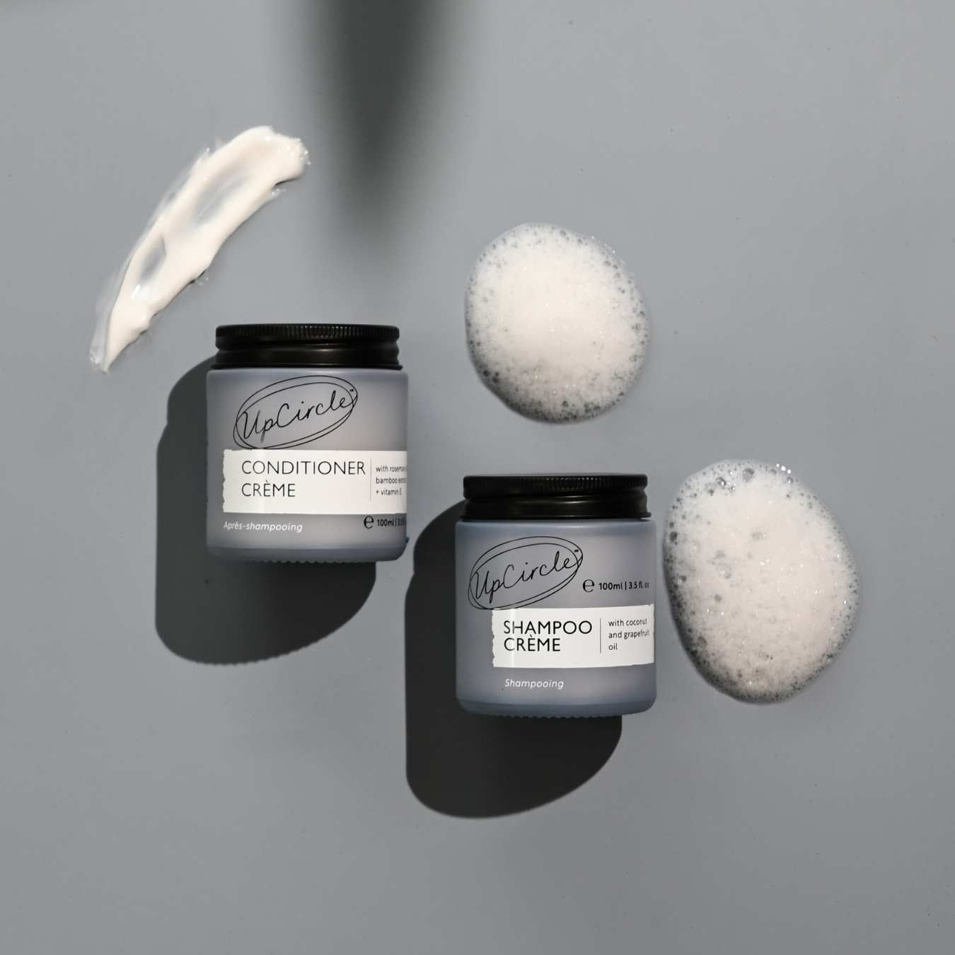 Two containers of UpCircle's Shampoo Creme and Conditioner Creme with applied cream on a gray background.