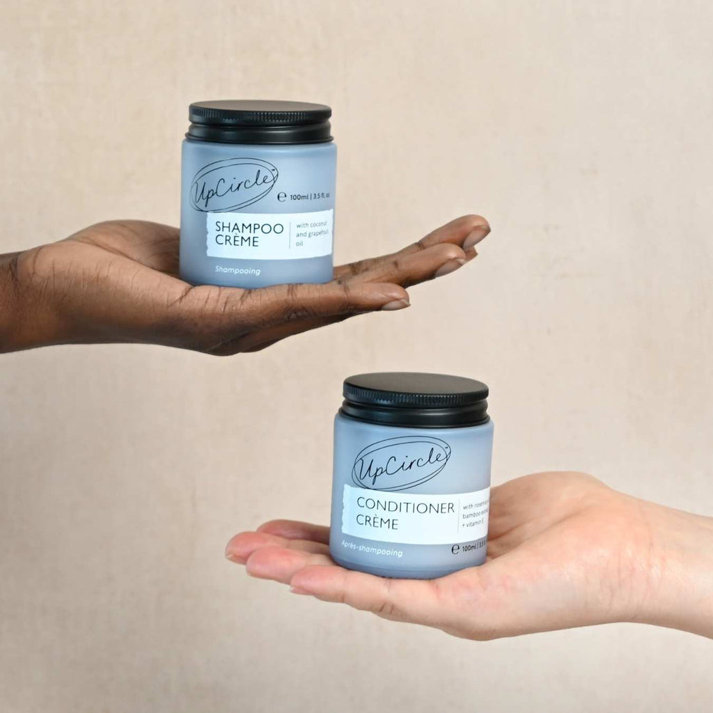 Two hands holding UpCircle's Shampoo Crème and Conditioner Crème jars against a beige background