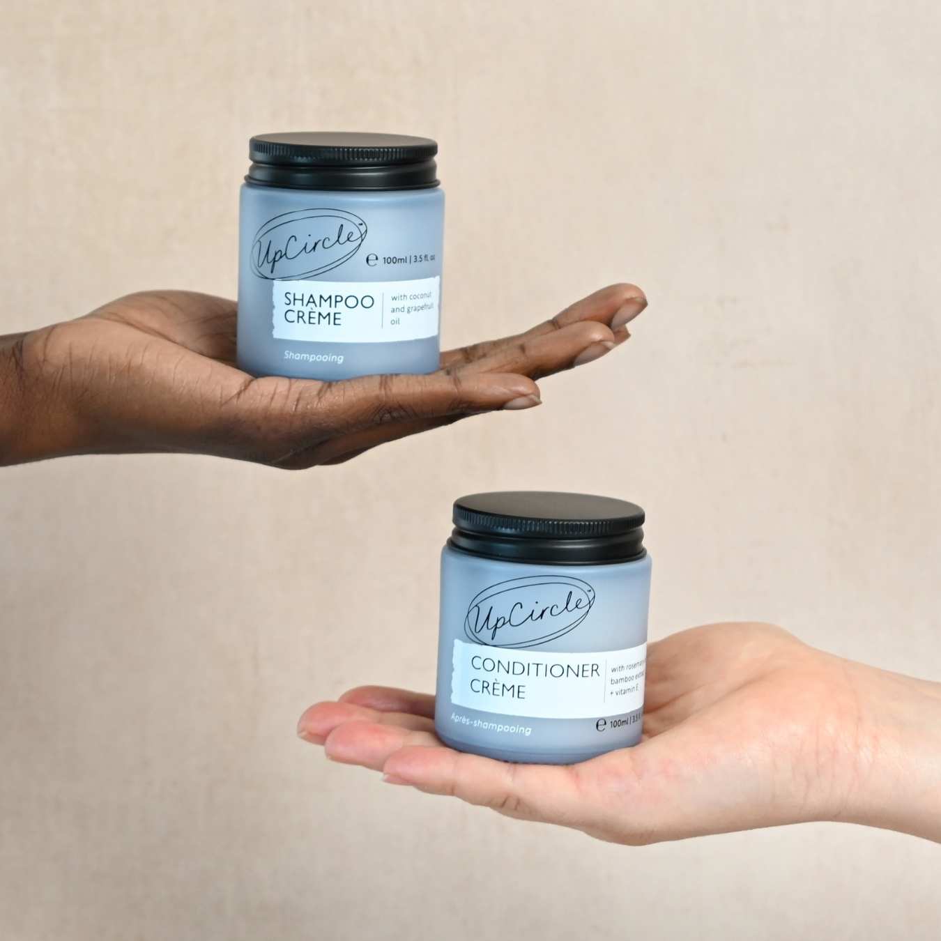 Two hands holding UpCircle's Shampoo Crème and Conditioner Crème jars against a beige background
