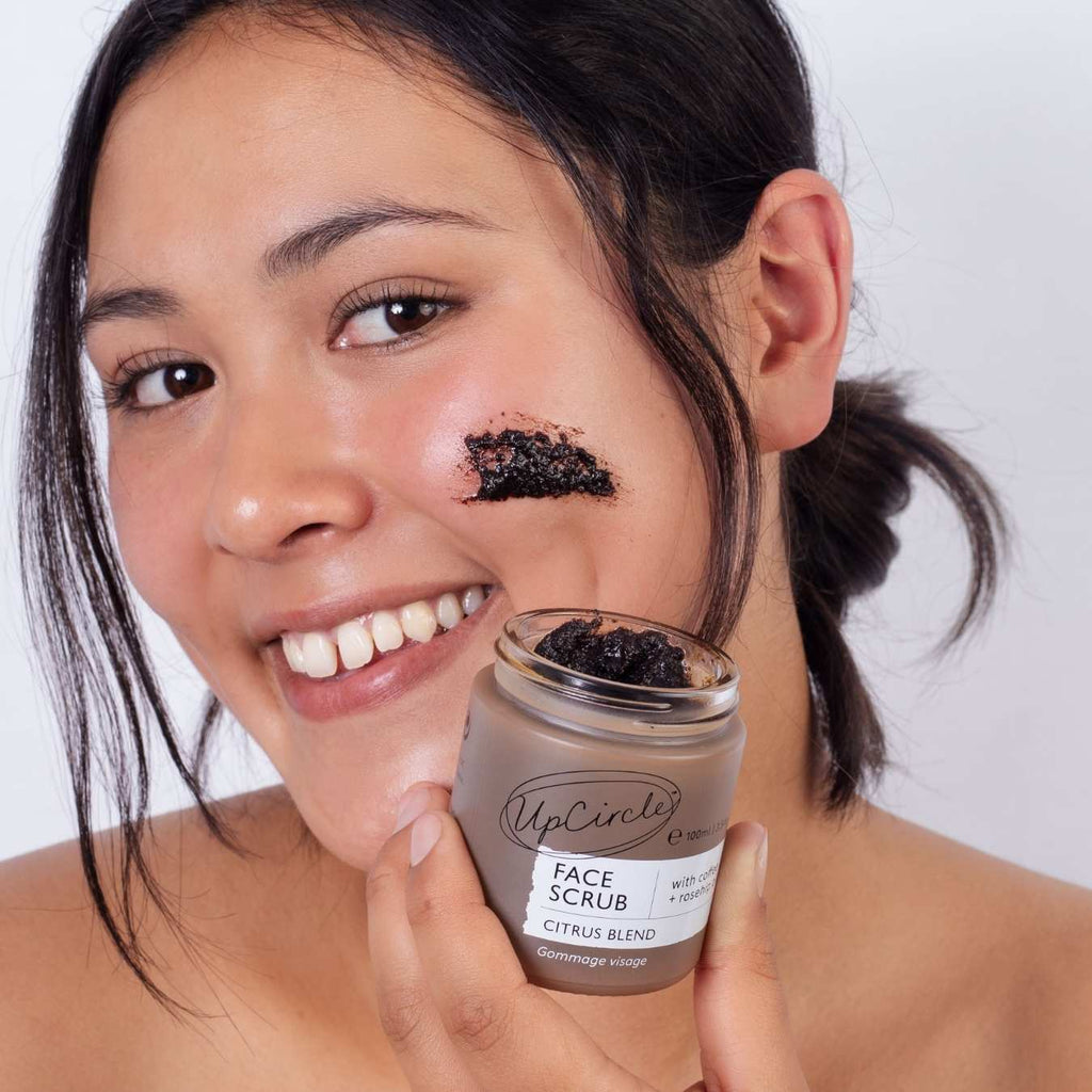 Woman with face scrub on cheek holding a jar of UpCircle Face Scrub.