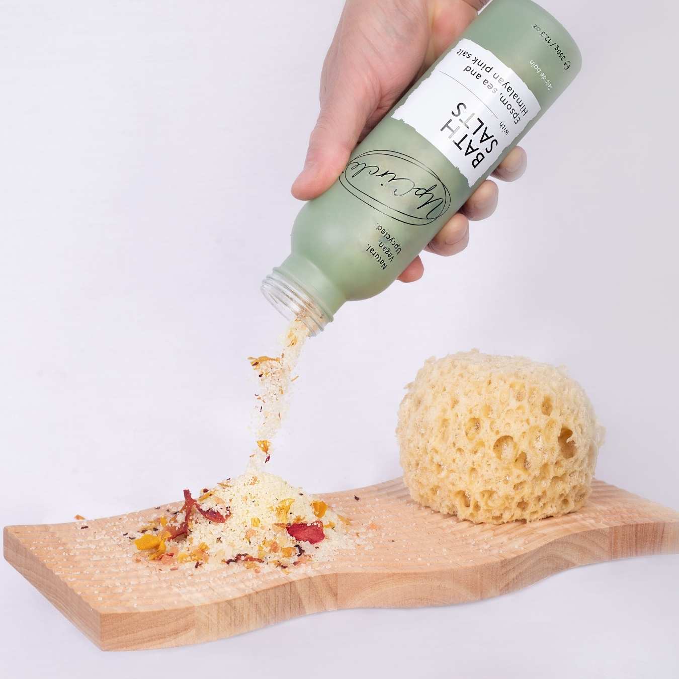 Hand holding a green bottle pouring bath salts onto a wooden board with a sponge