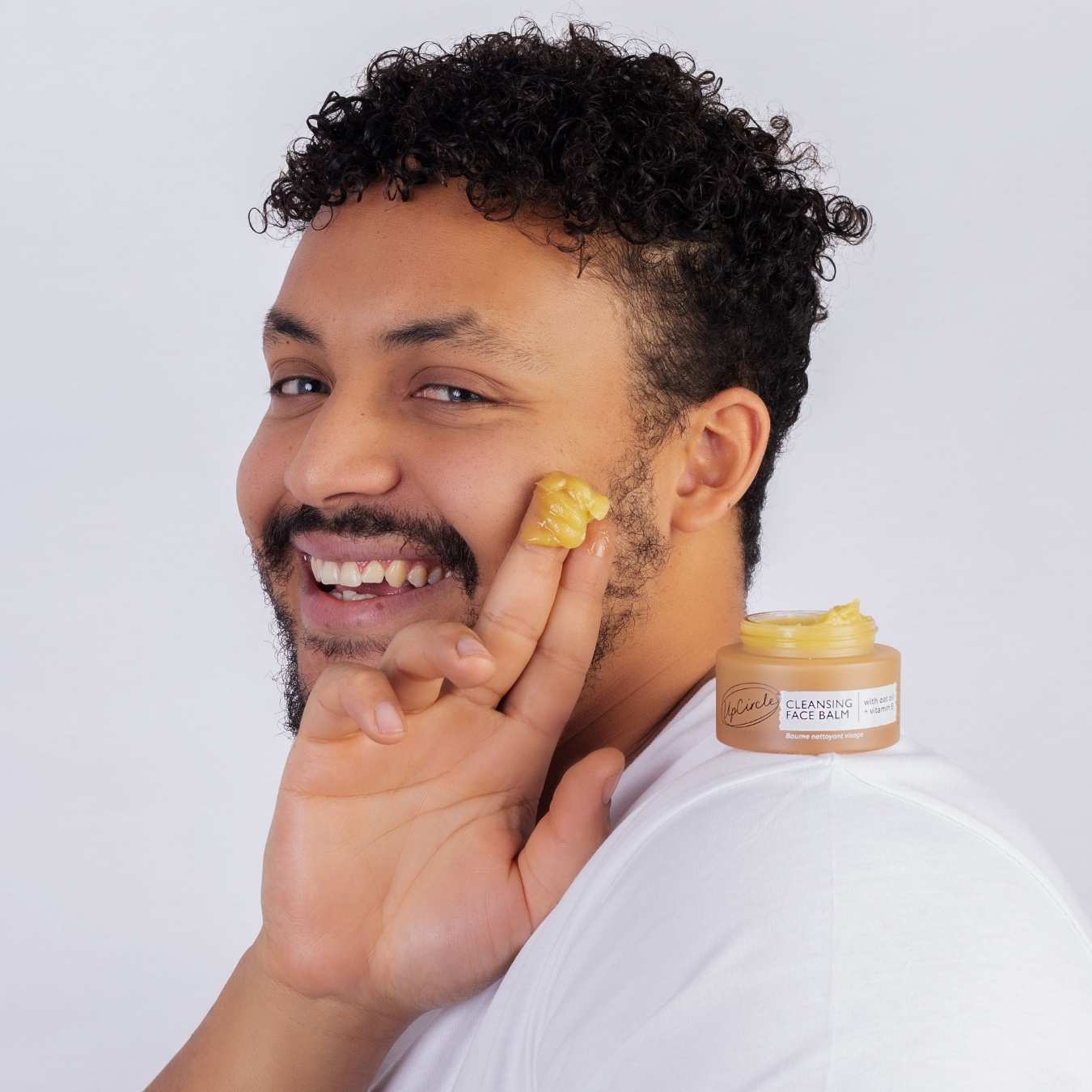 Man applying yellow cream to his face with a jar of the same cream on a white background