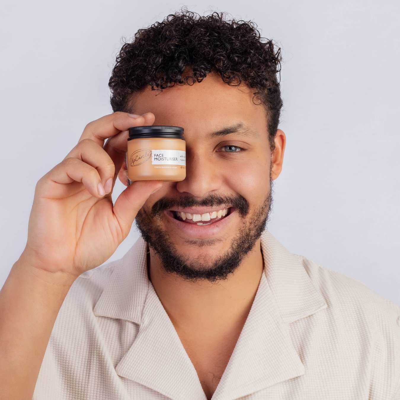 Man holding a jar of skin product in front of his face against a plain background