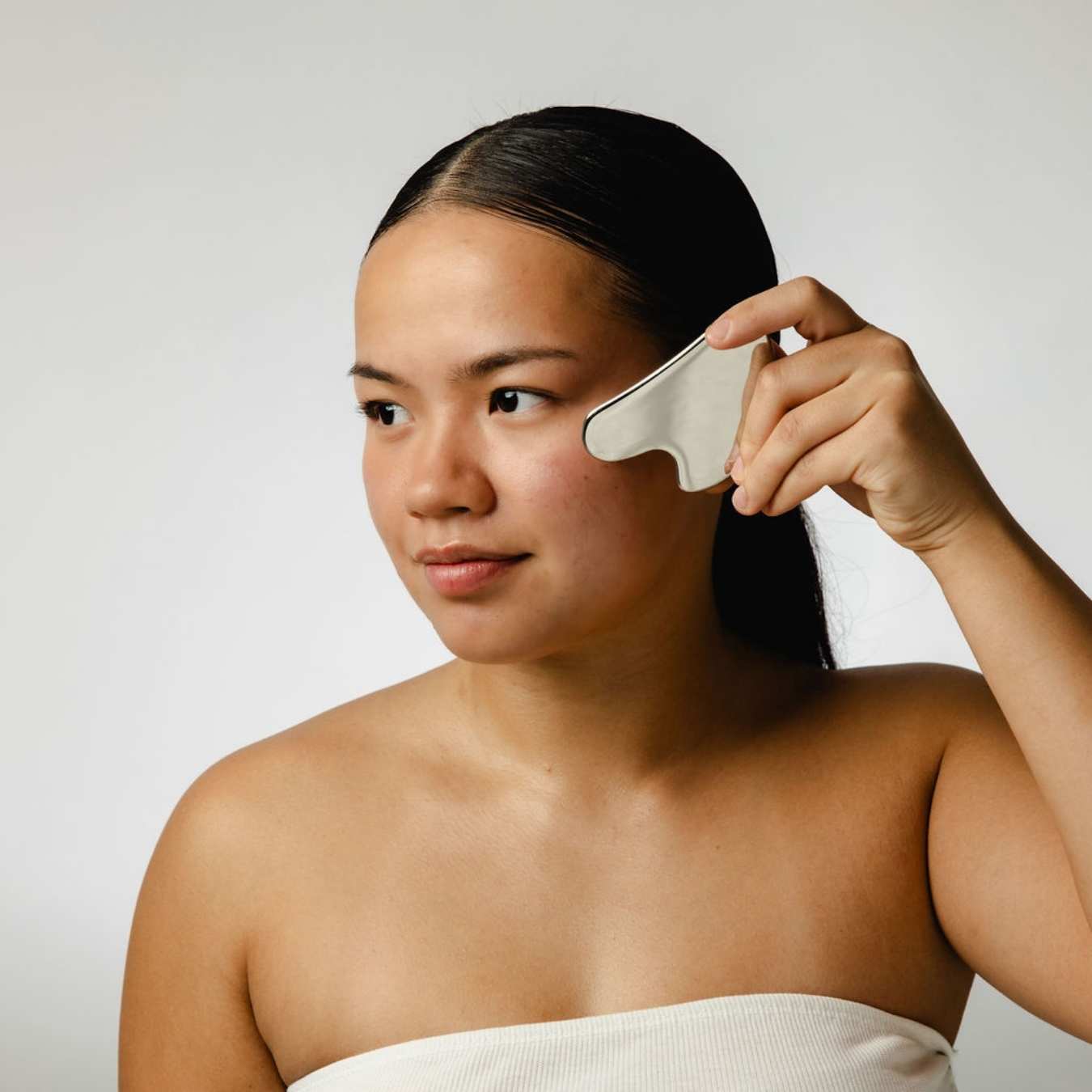 Woman using a silver gua sha tool on her face against a plain background