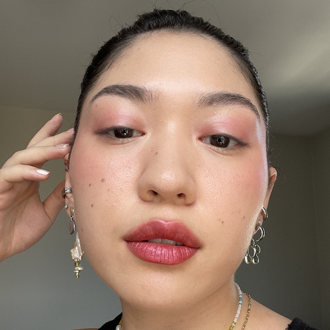 Close-up of a person with makeup, wearing earrings and a necklace, against a neutral background.