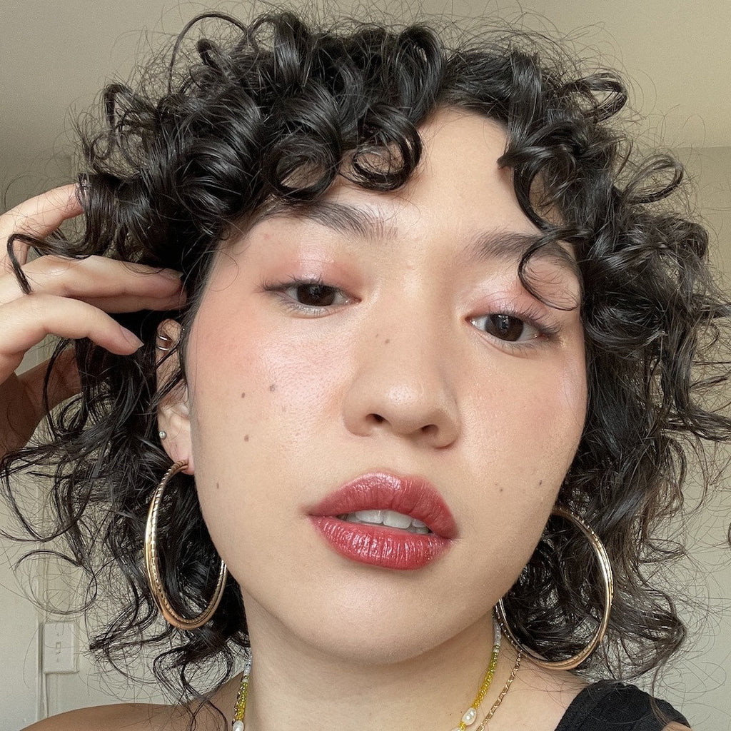 Close-up of a person wearing makeup with curly hair and hoop earrings, wearing a black top.