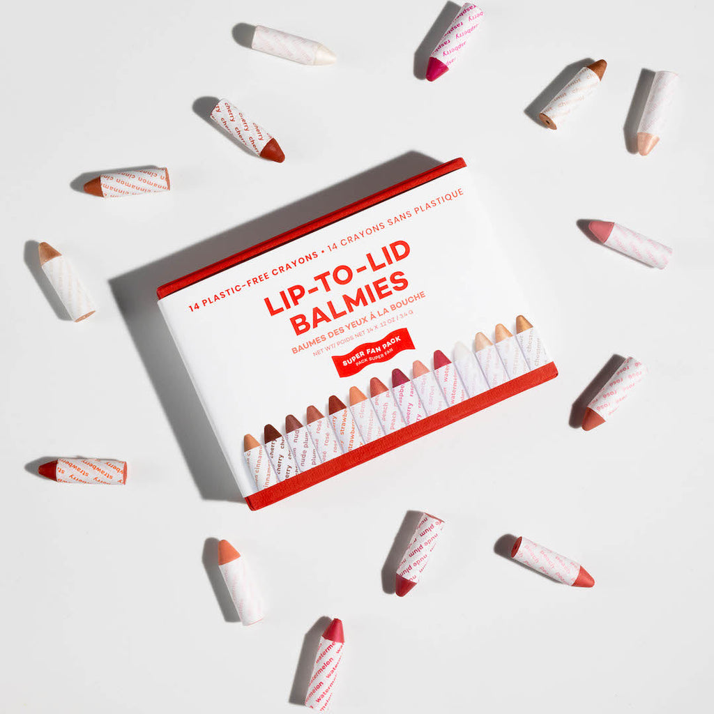 Packaging of Lip-To-Lid Balmies with lip balm sticks scattered around on a light gray background