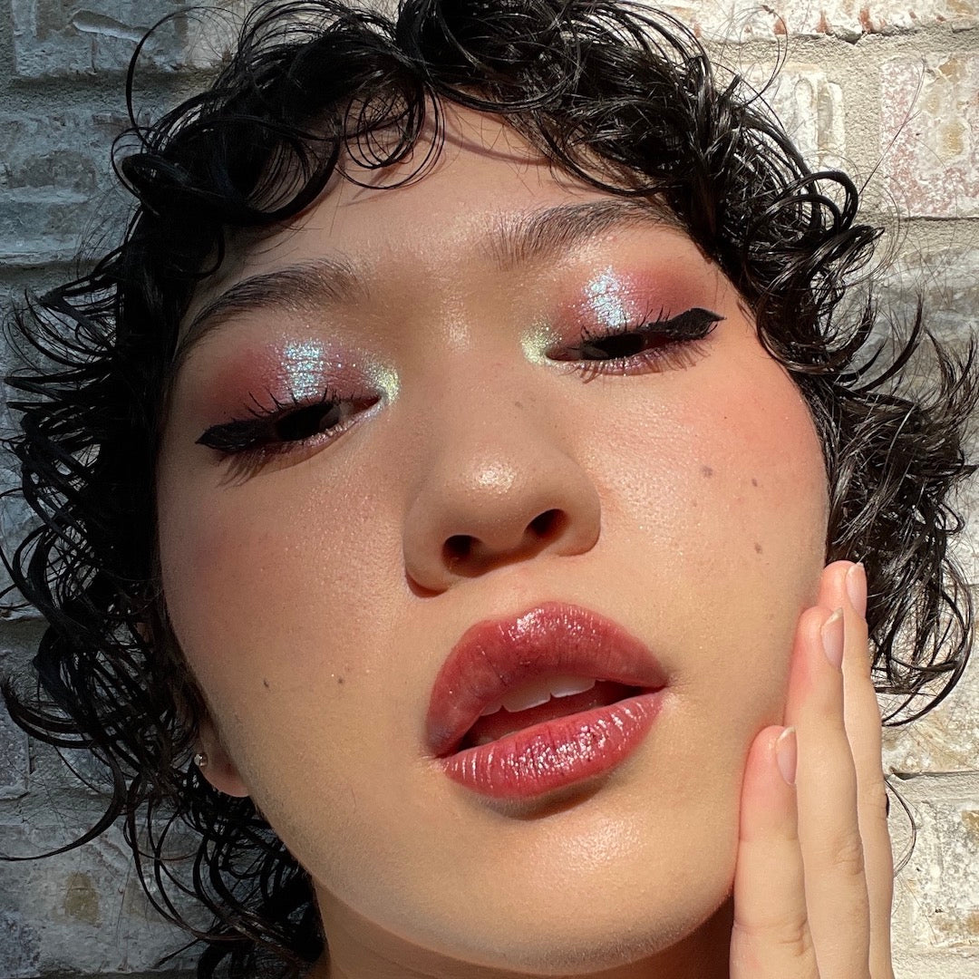 Close-up of a person with glittery eye makeup and pink lipstick against a stone wall background.