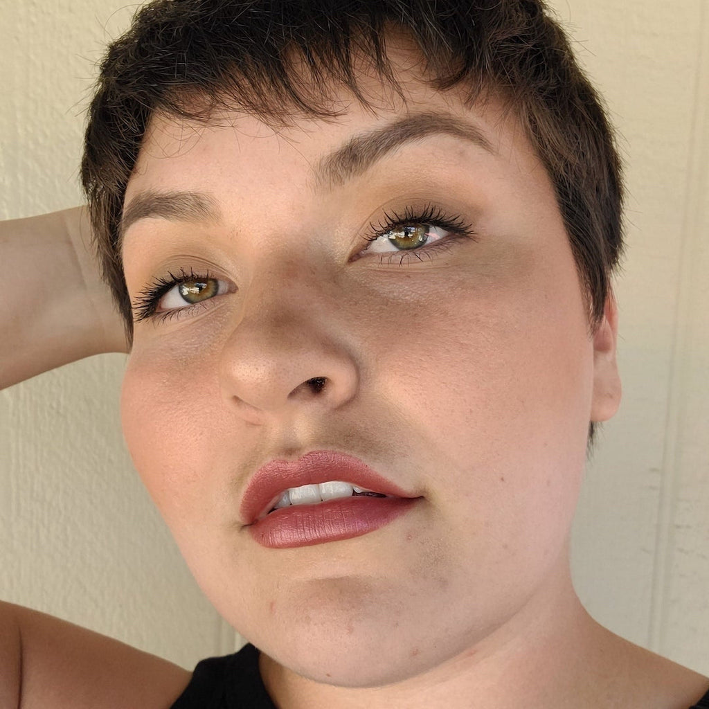 Close-up of a person with short hair and neutral expression wearing makeup against a plain background