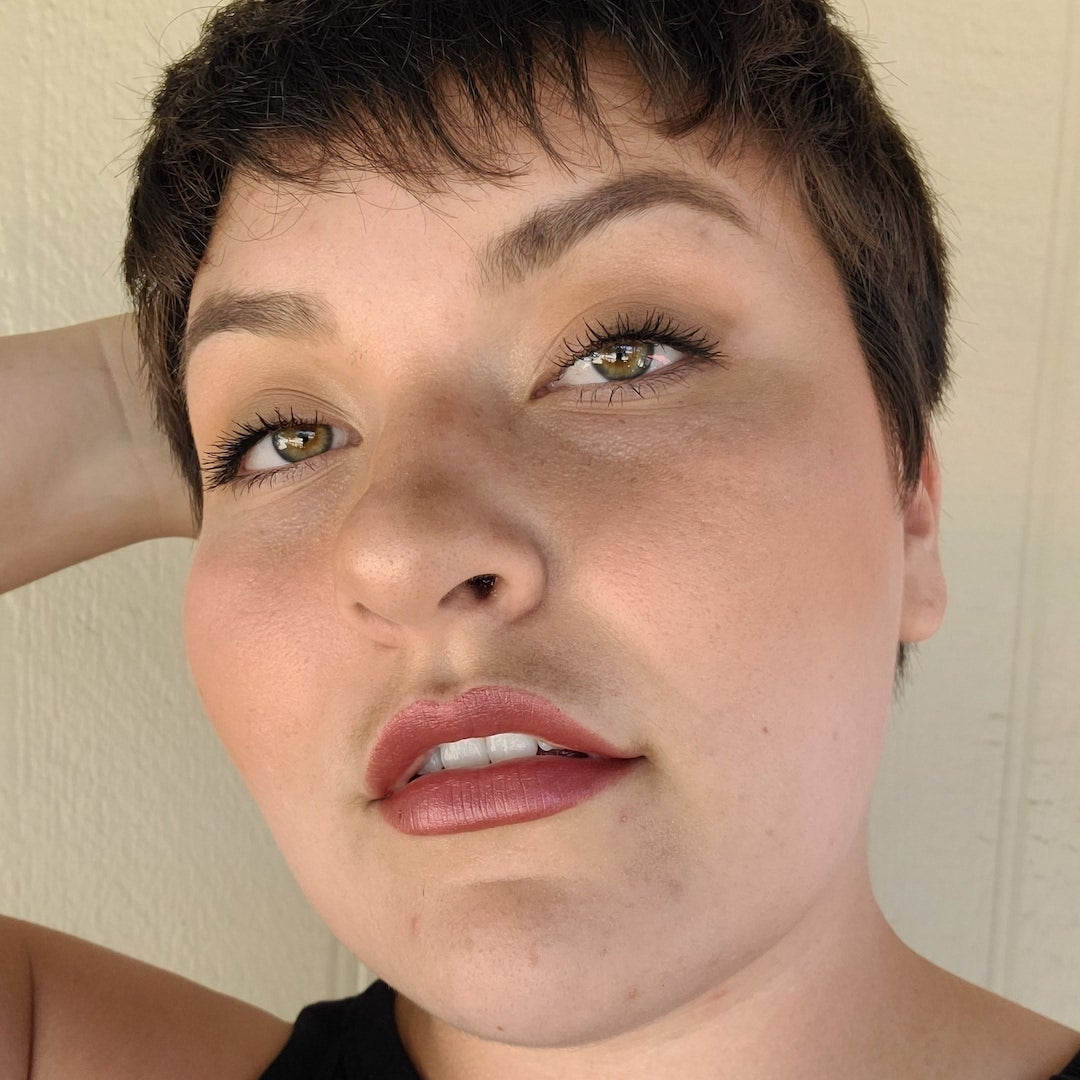 Close-up of a person with short hair and neutral expression wearing makeup against a plain background