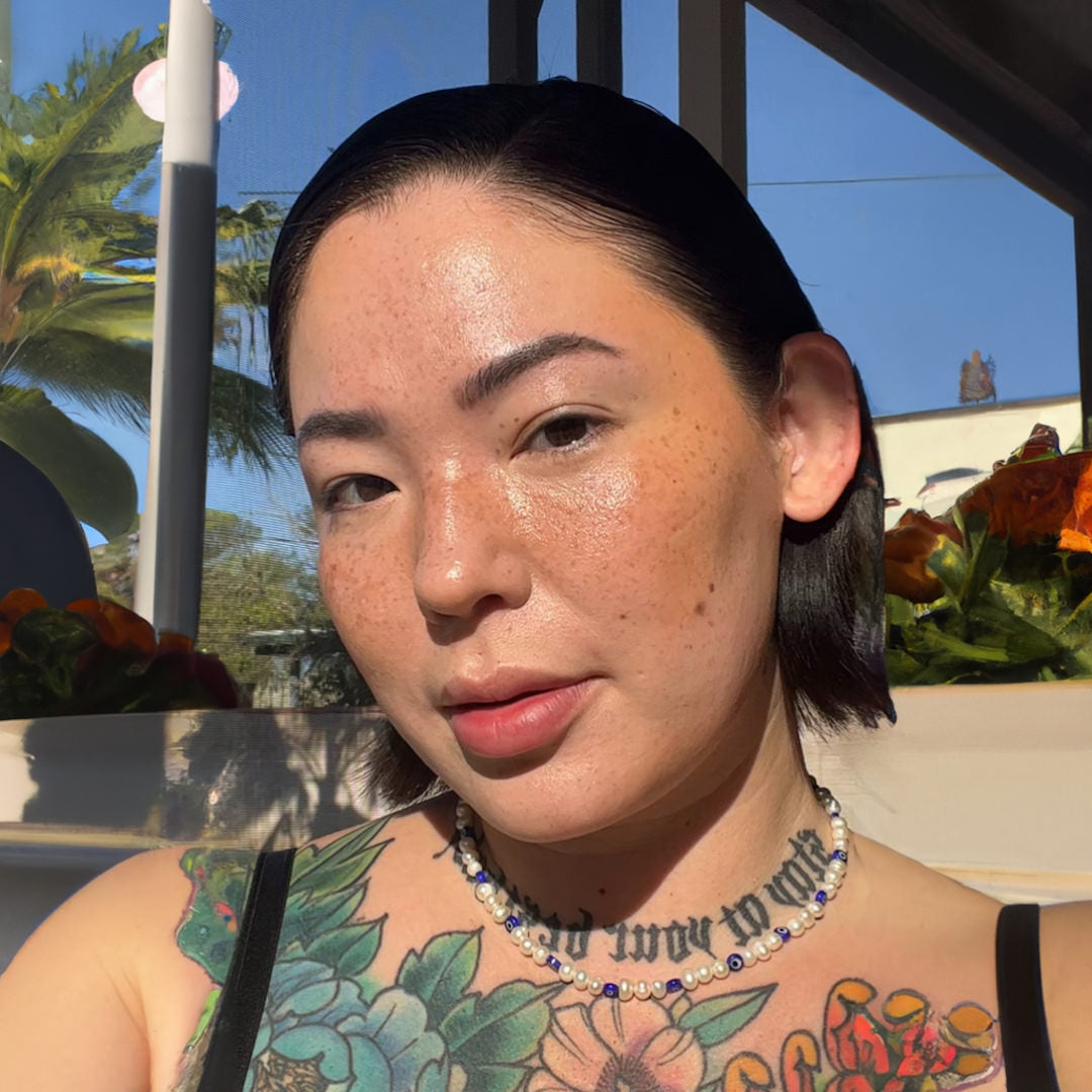 Person with tattoos and a necklace standing outdoors with plants in the background