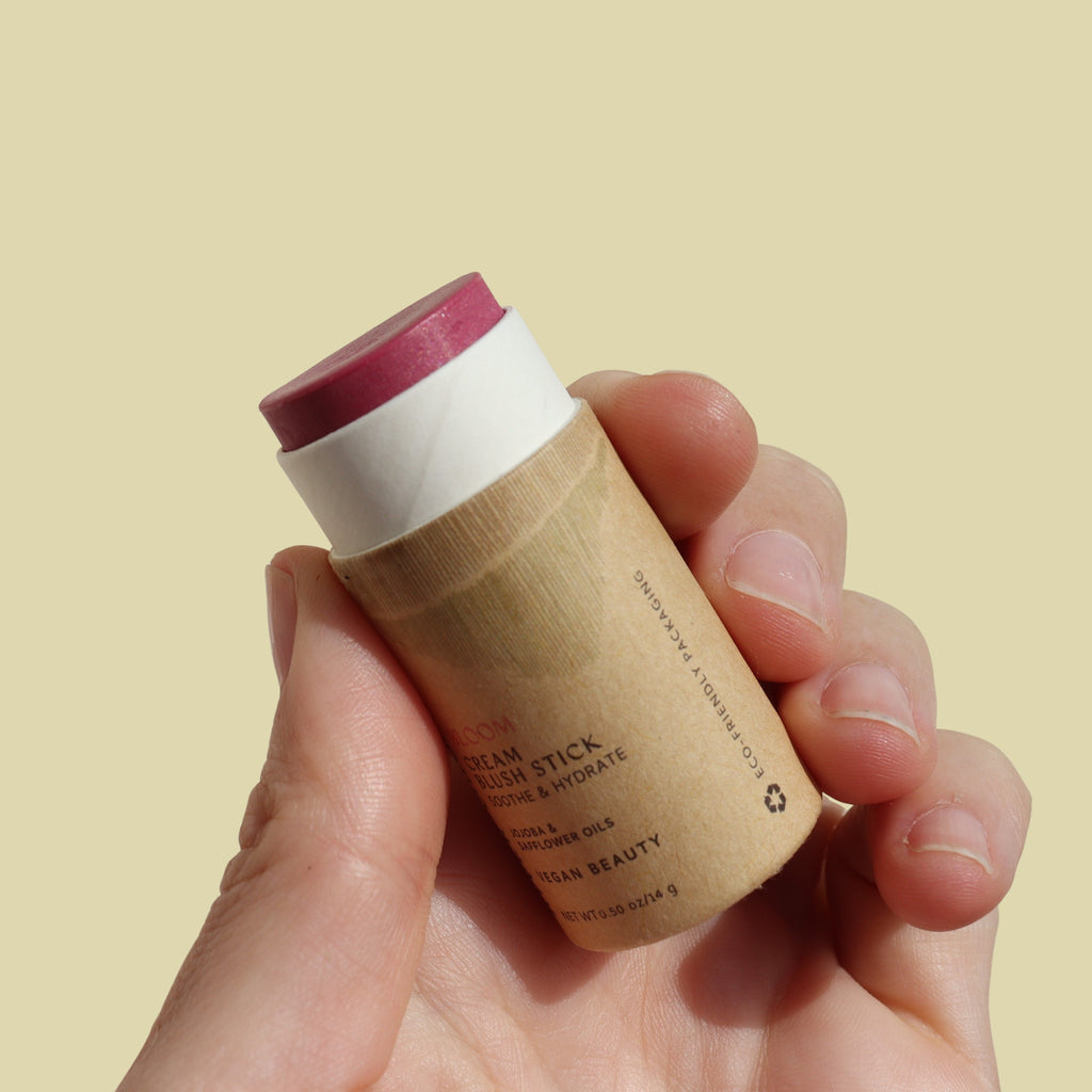 Hand holding a cylindrical cream blush stick with a beige background