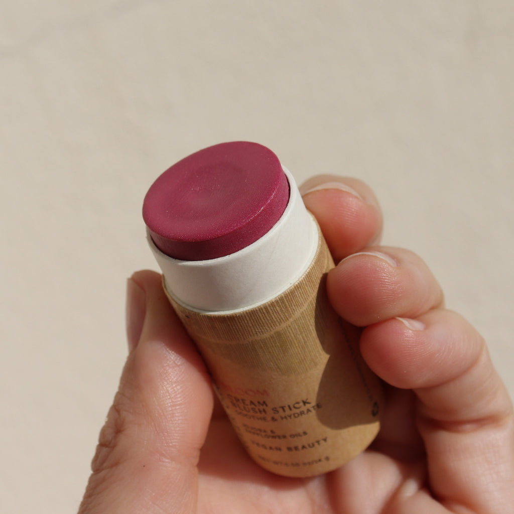Hand holding a blush stick in paper tube against a neutral background