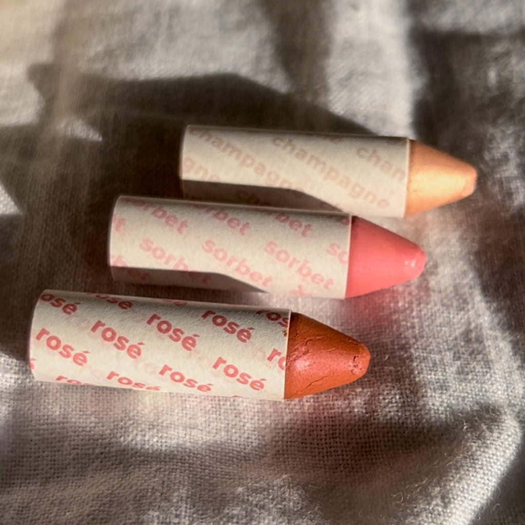 Three makeup crayons with 'champagne', 'sorbet', and 'rosé' text on their wrappers, lying on a textured surface.
