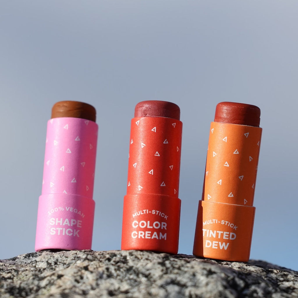 Three cosmetic sticks in pink, red, and orange on a rock with a clear sky background