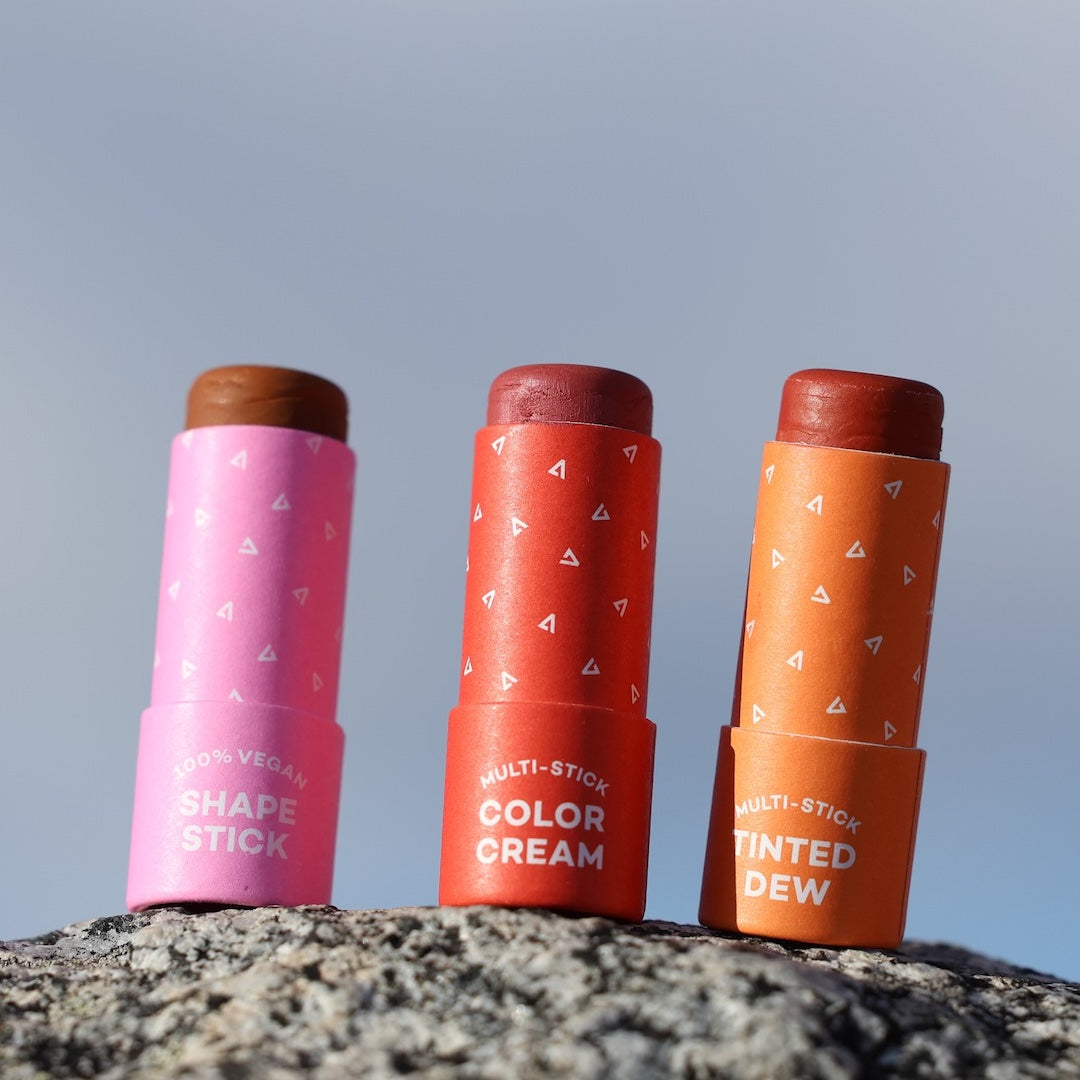 Three cosmetic sticks in pink, red, and orange on a rock with a clear sky background