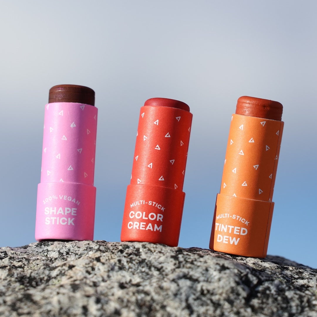 Three cosmetic sticks in pink, red, and orange on a rock with a blue sky background