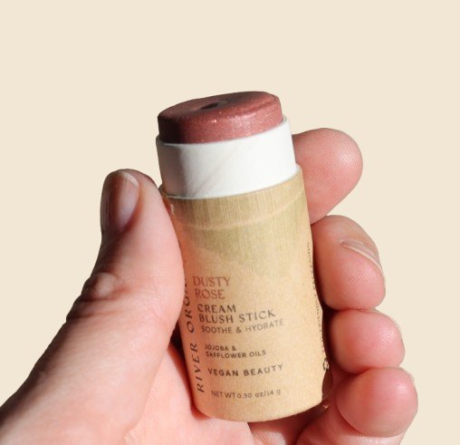 Hand holding a River Organics blush stick against a beige background