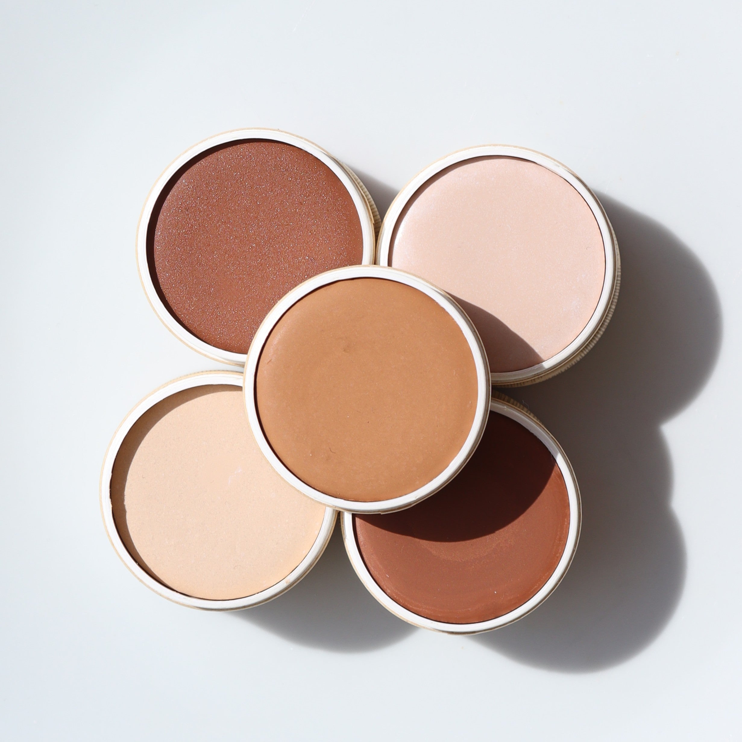 Five round concealer pots in various shades of brown and beige on a light gray background
