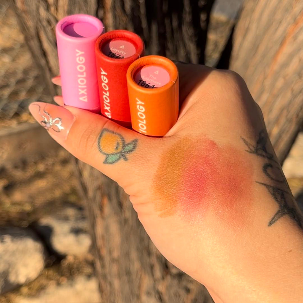 Three cosmetic tubes from the brand 'Axiology' held by a hand with makeup swatches on the skin.