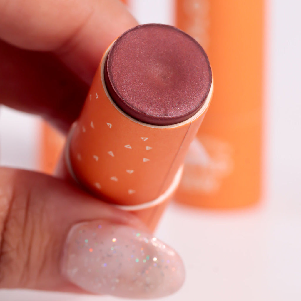 Lipstick with a brown shade held by a hand against an orange background
