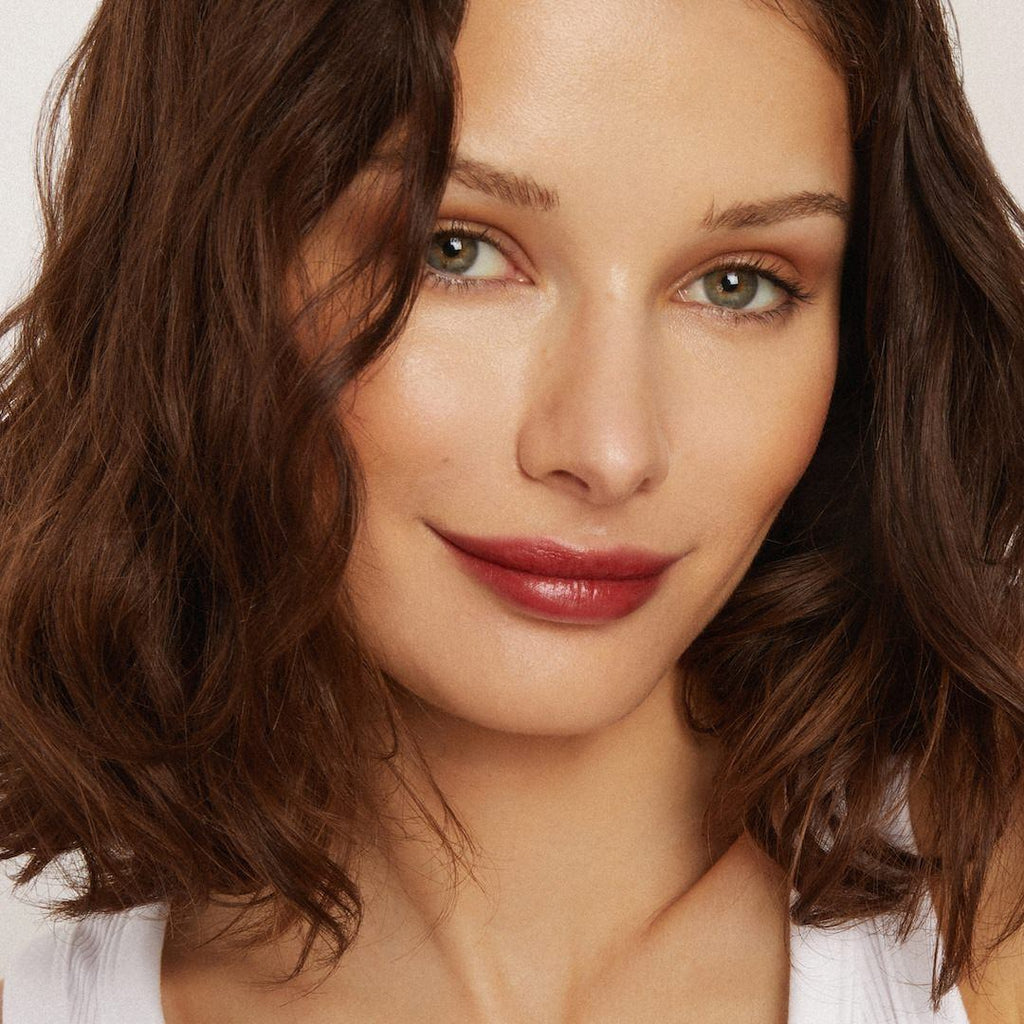 Close-up of a woman with wavy brown hair wearing foundation makeup and red lipstick on a plain background