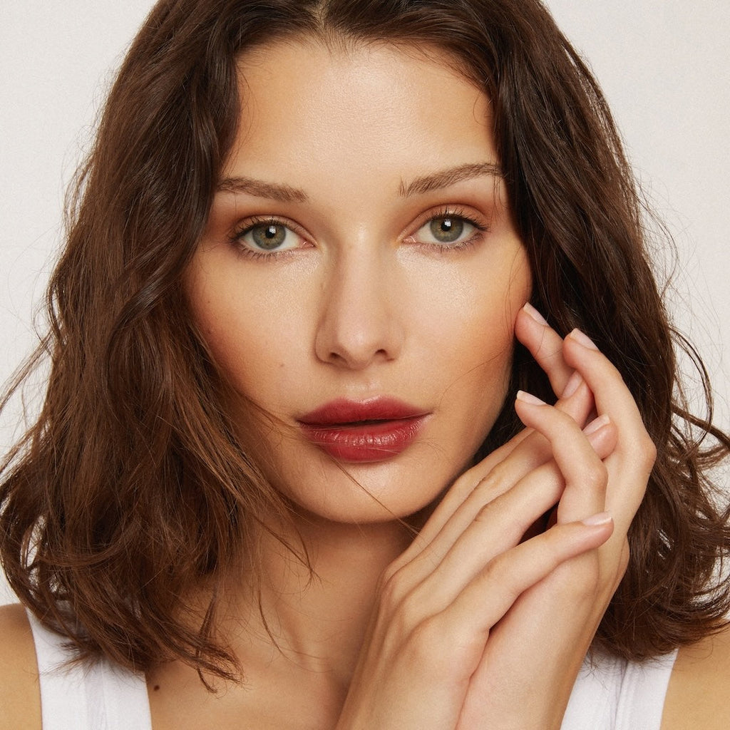 Woman with brown hair and red lipstick touching her face against a neutral background