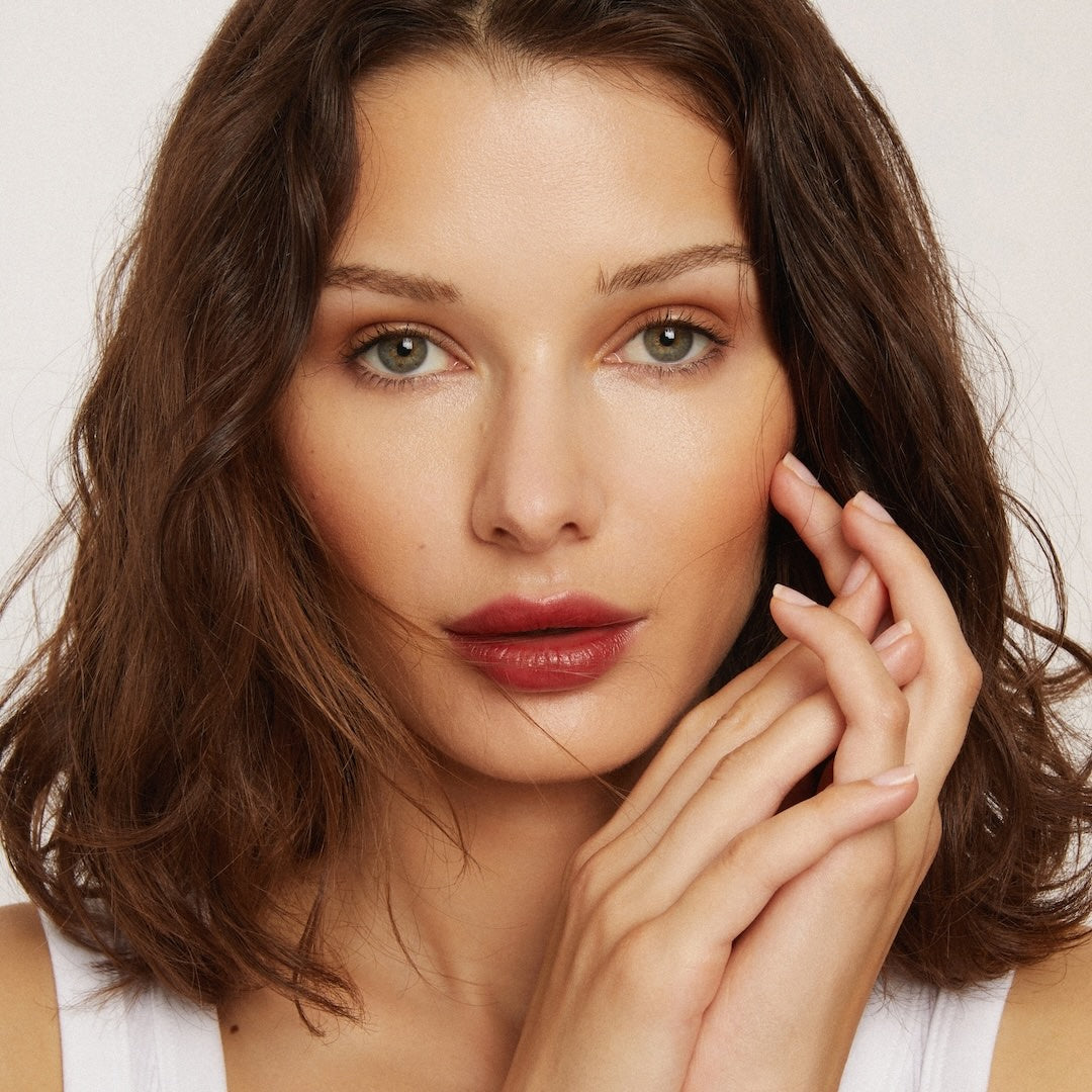 Woman with brown hair and red lipstick touching her face against a neutral background