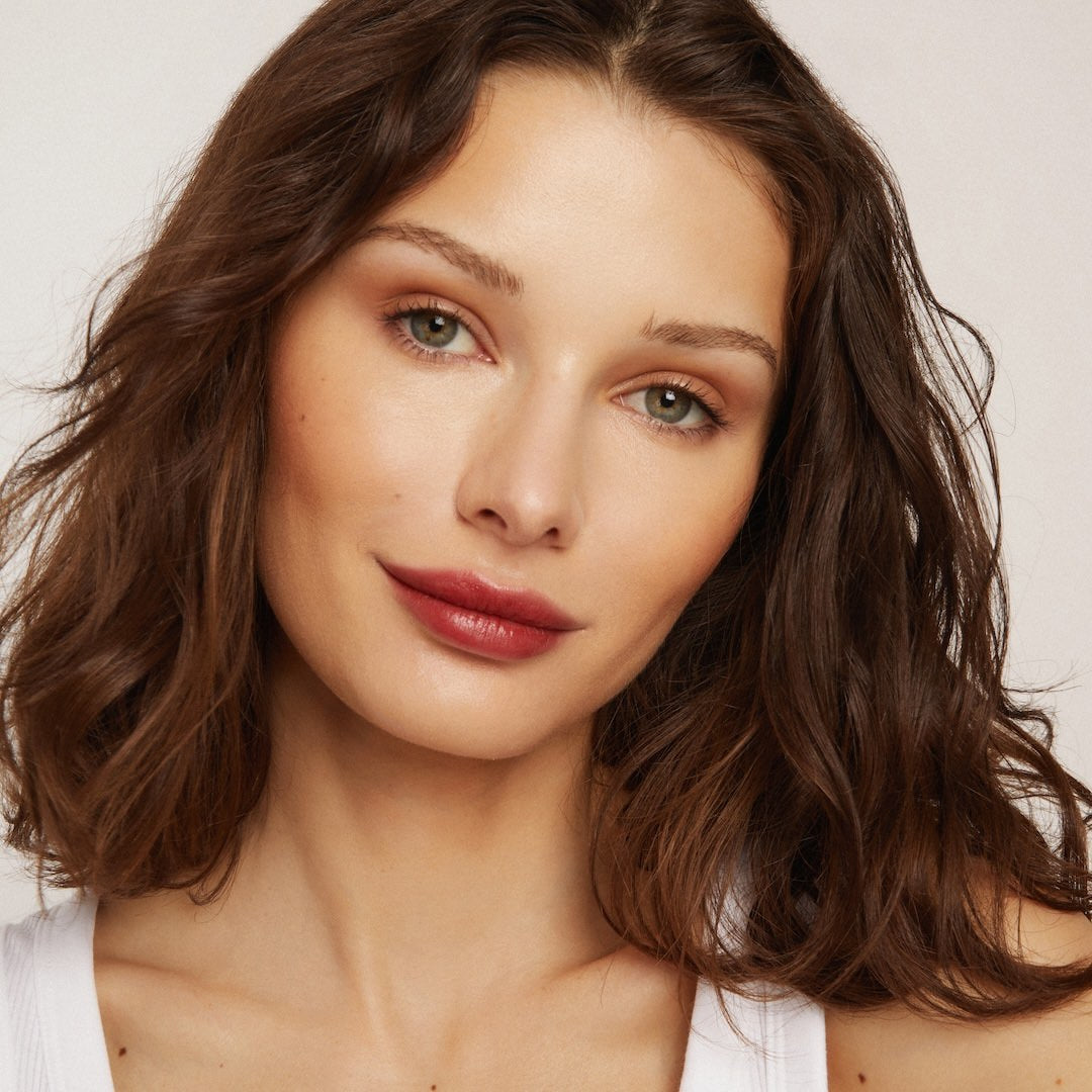 Woman with wavy brown hair wearing foundation makeup and red lipstick against a neutral background