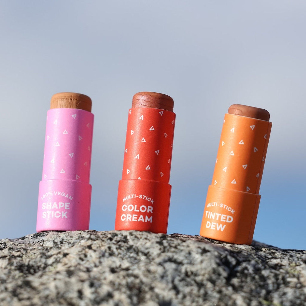Three cosmetic sticks in pink, red, and orange on a rock with a blue sky background