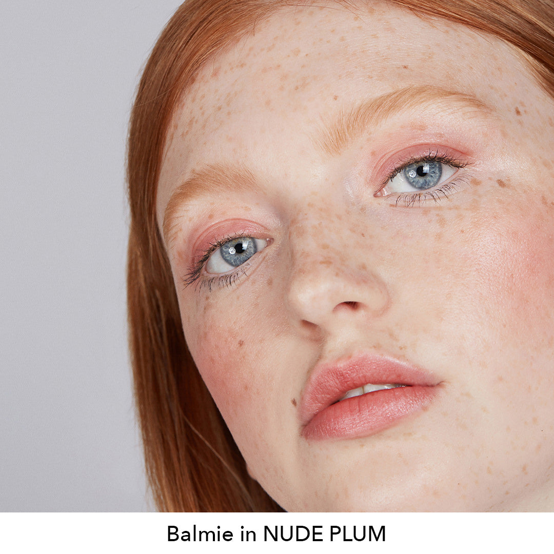 Close-up of a person with red hair and freckles, wearing makeup with 'Balmie in NUDE PLUM' text.