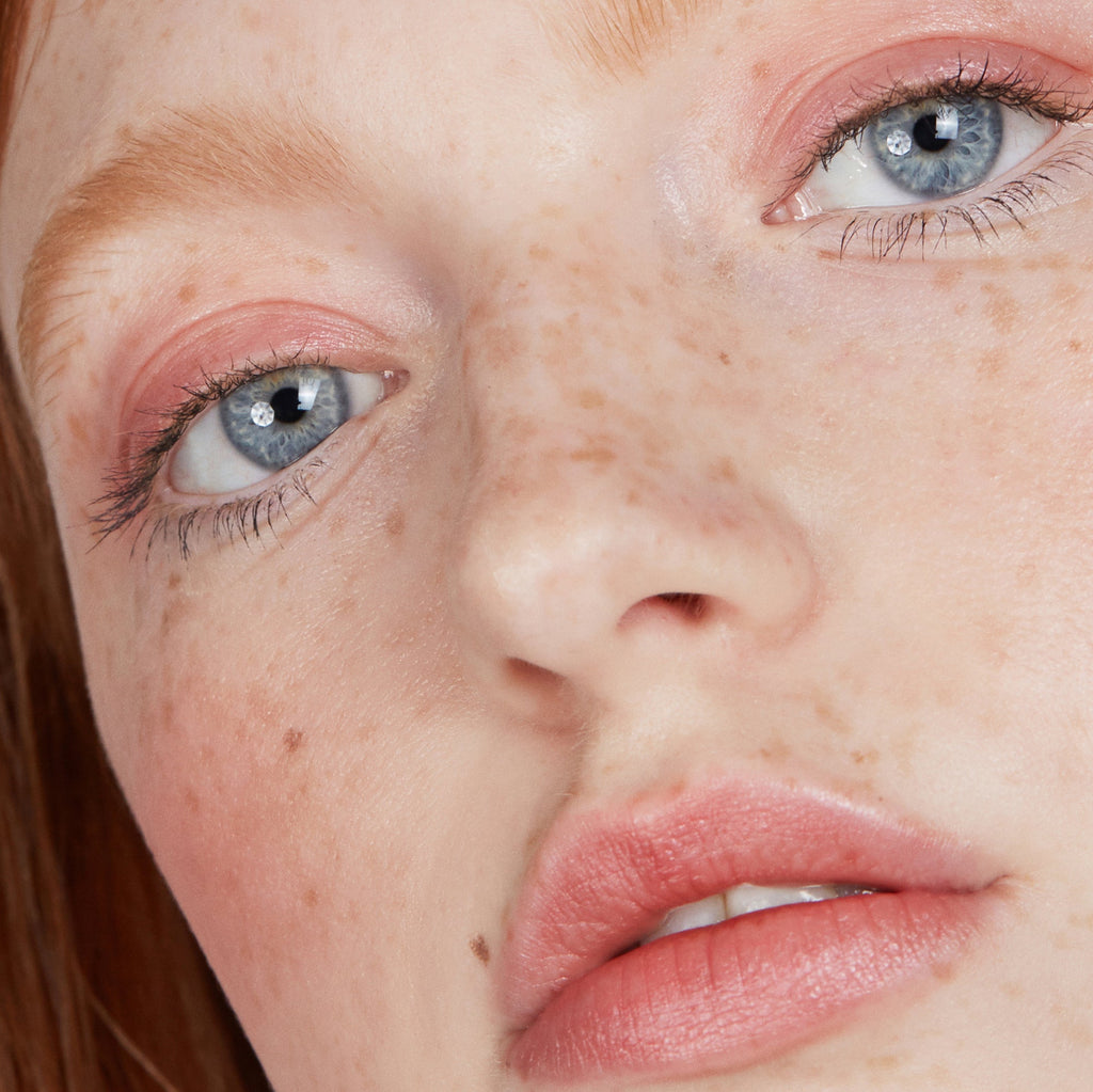 Close-up of a young woman's face with freckles and blue eyes, wearing makeup.