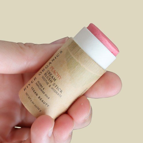 Hand holding a cream blush stick with a beige background