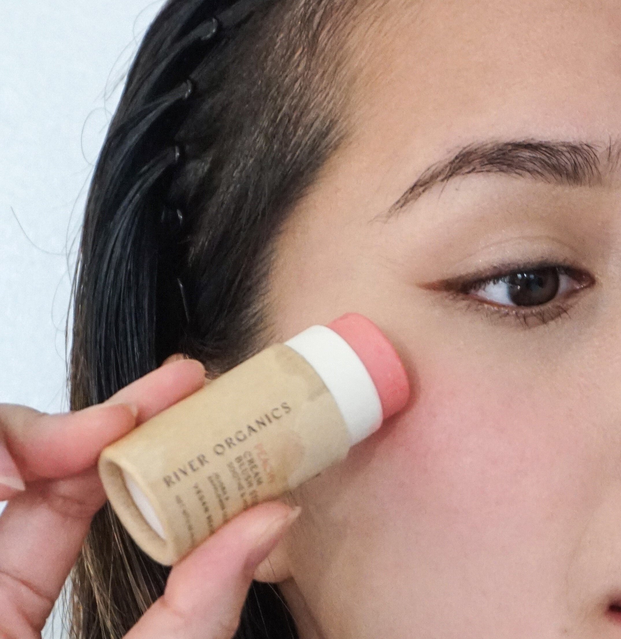 Person applying a stick of 'River Organics' cream blush to their face.