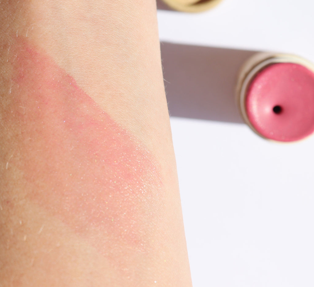 Pink blush swatch on a person's arm with the blush stick visible in the background.