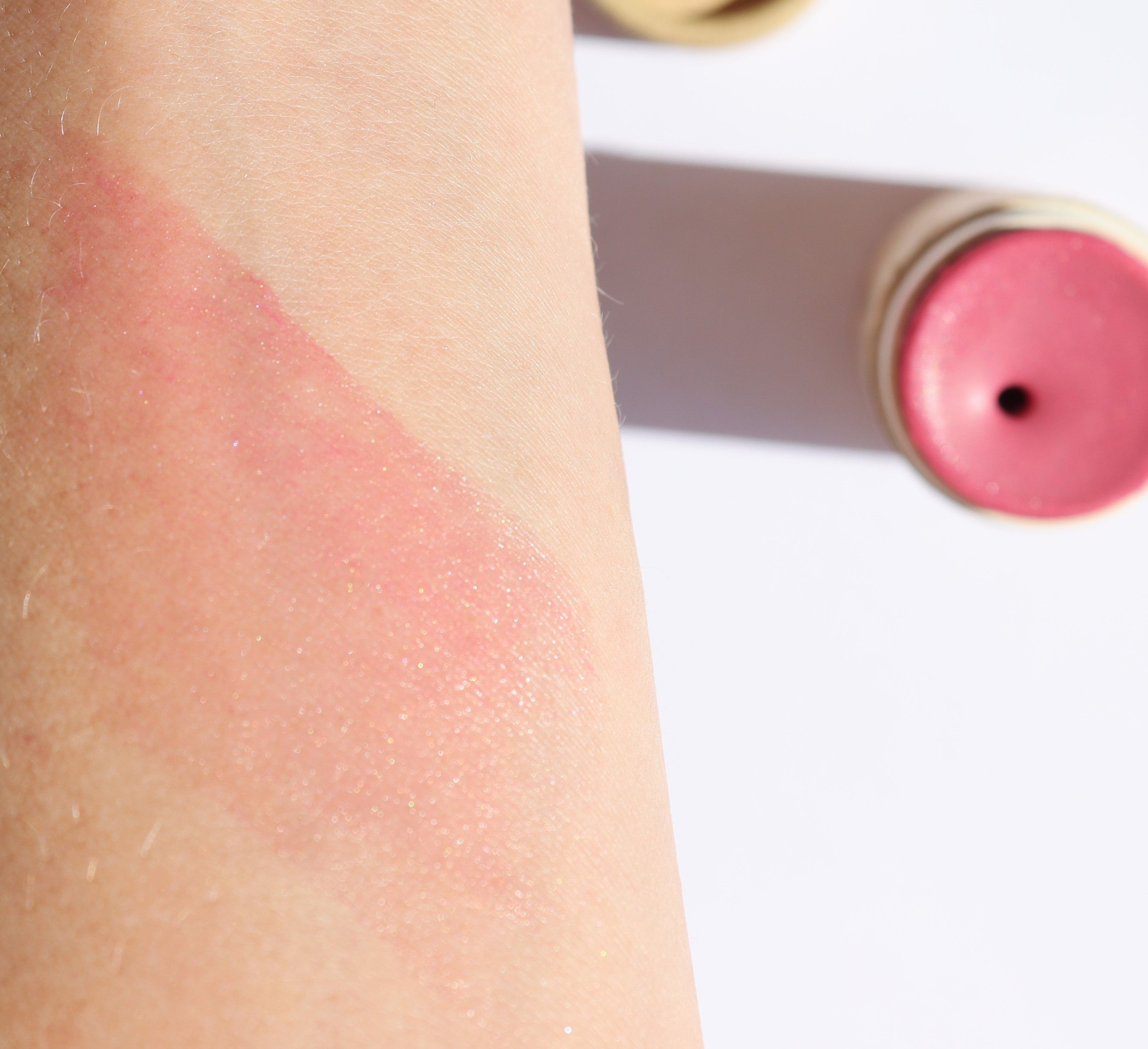 Pink blush swatch on a person's arm with the blush stick visible in the background.