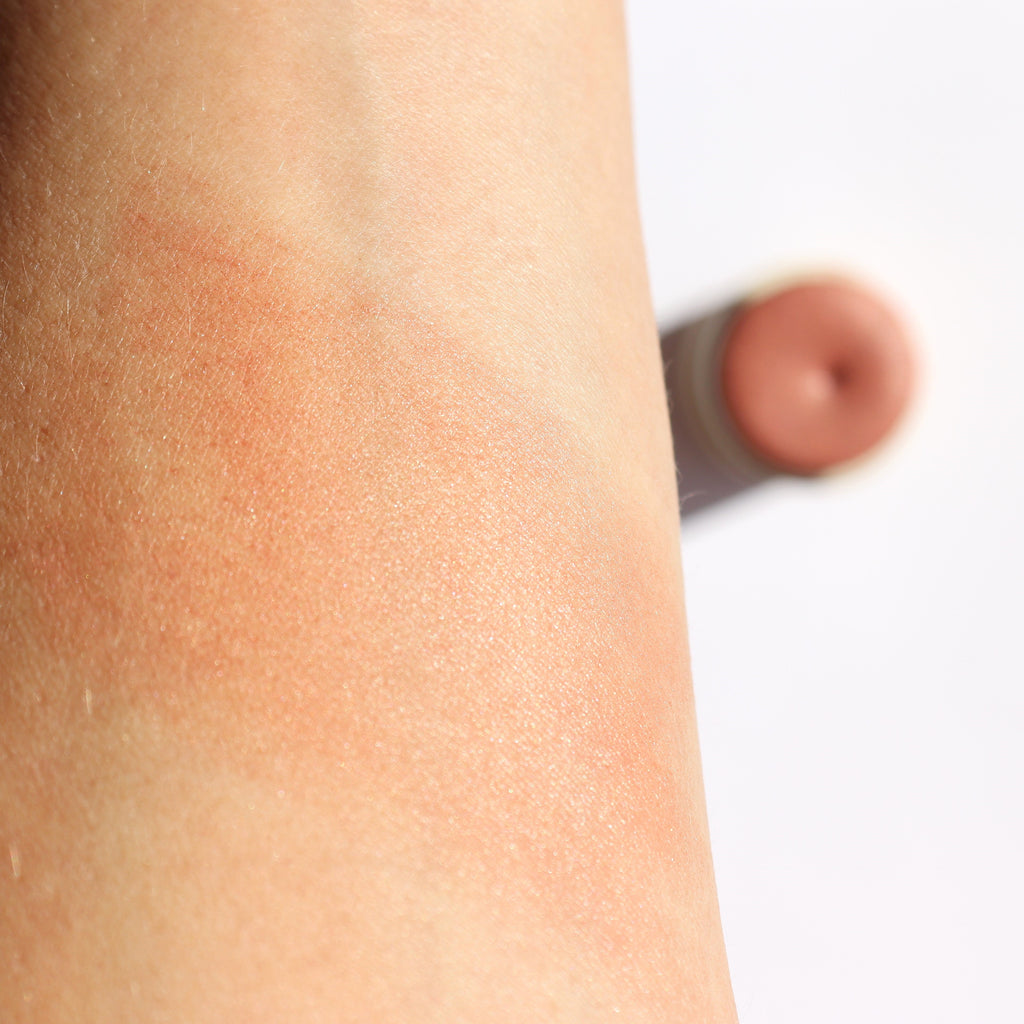Close-up of a swatch of blush on skin with a blurred blush stick in the background