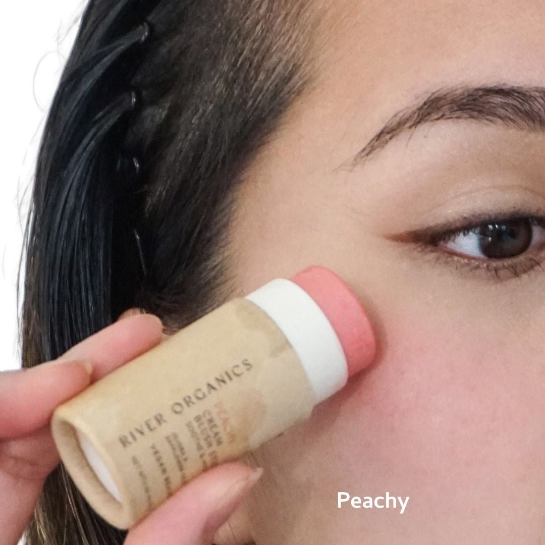 Person applying a Peachy-colored blush stick to their face with 'River Organics' branding.