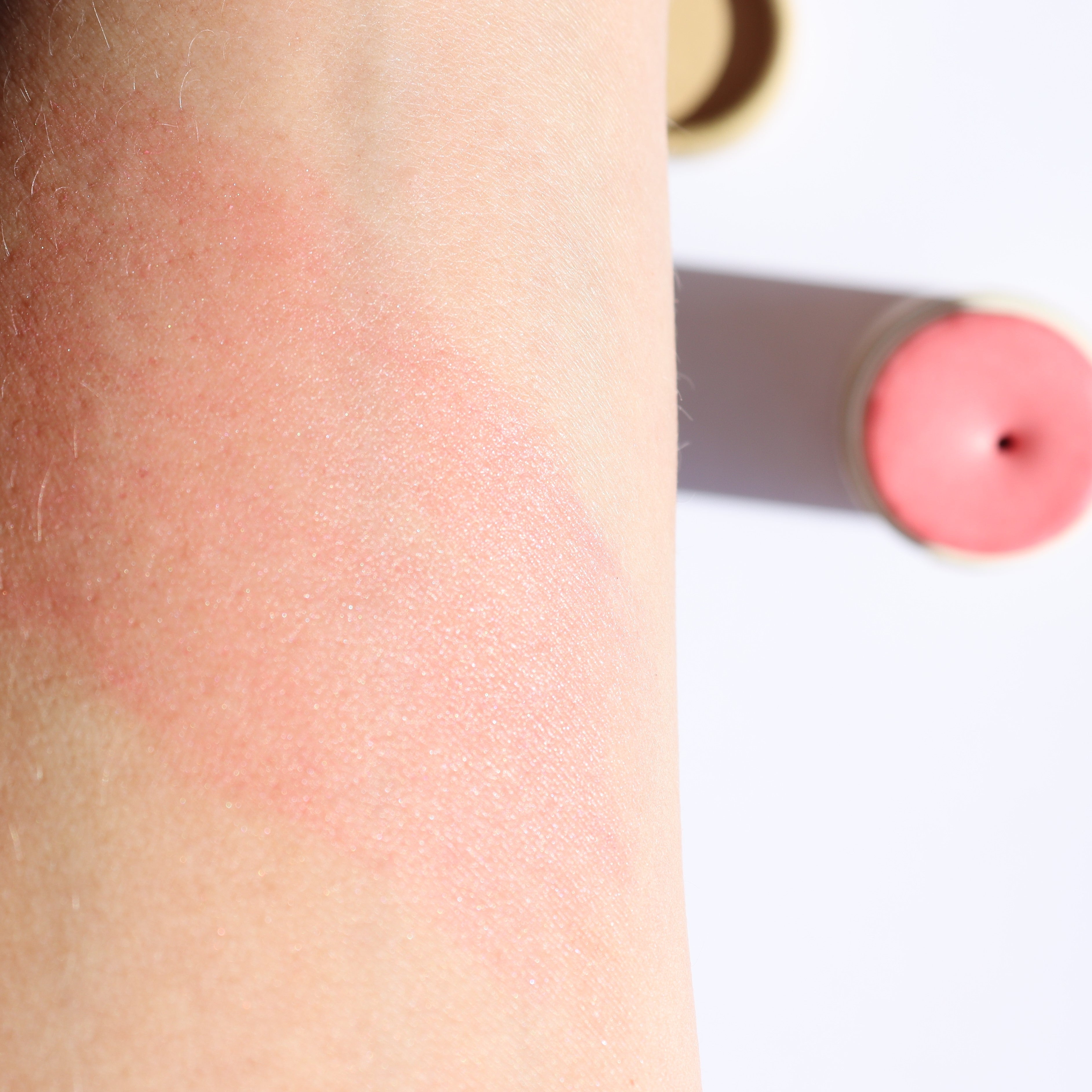Close-up of a pink blush swatch on skin and a tube of blush in the background.