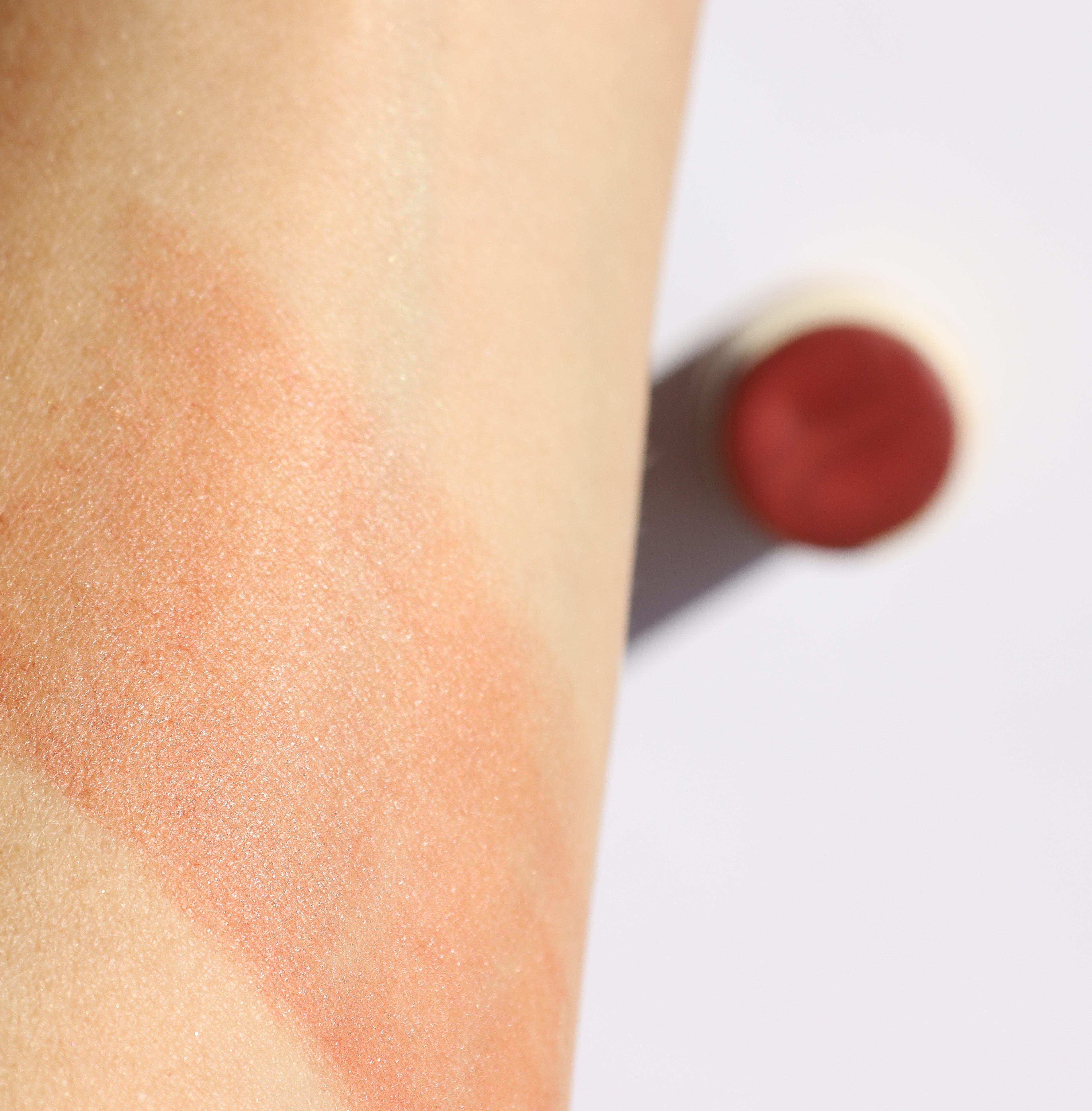 Close-up of a swatch of makeup on skin with a blurred blush stick in the background.