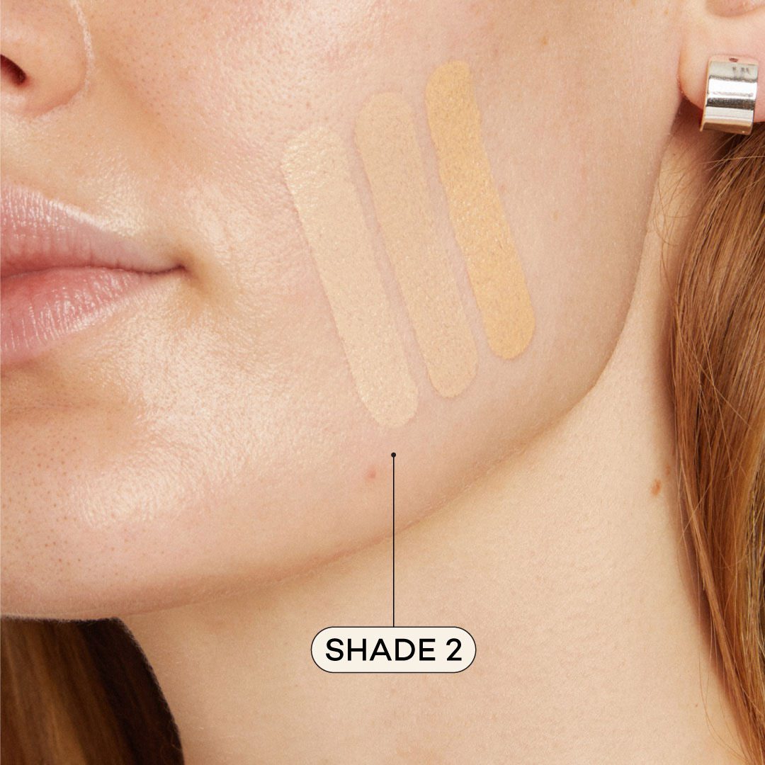 Close-up of a woman's face with foundation swatches on her cheek, labeled 'Shade 2'.