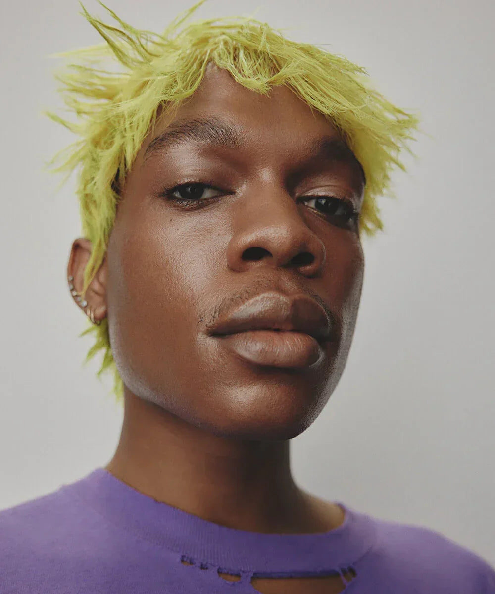 Person with bright yellow hair wearing highlighter makeup and a purple shirt against a plain background