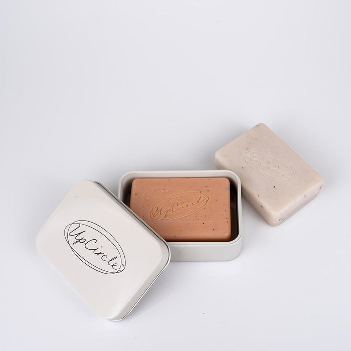 Brown bar of soap in a white tin with 'UpCircle' branding on a light gray background